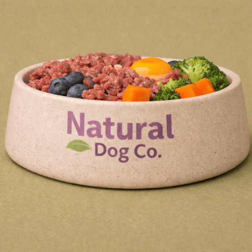 Front view of a Natural Dog Co branded bowl filled with raw meat, blueberries, carrots, broccoli and a whole raw egg with shell, showing a nutritionally enriched raw dog food meal
