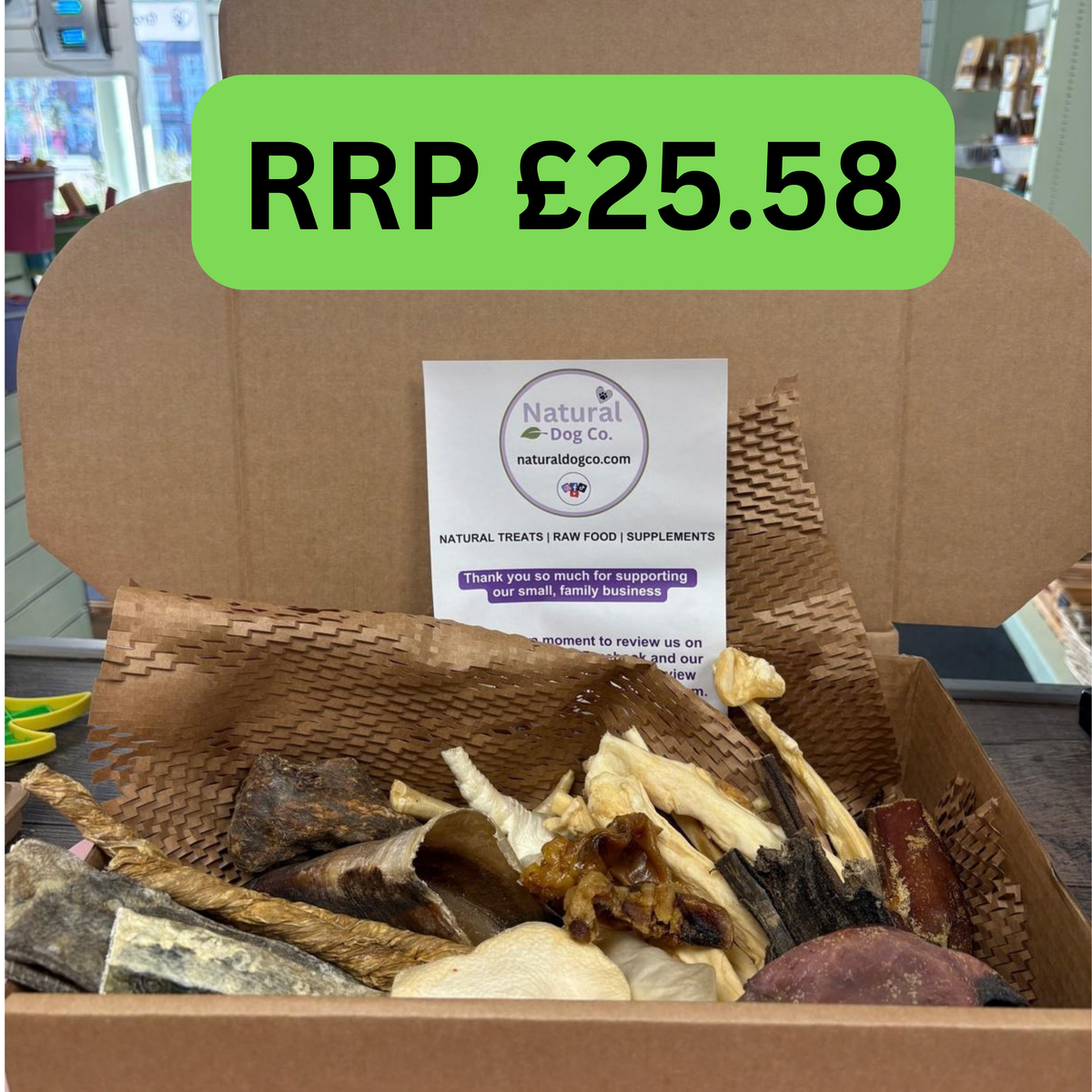 An open cardboard box filled with dried dog treats and a branded card from Natural Dog Co., with a bright green overlay reading 'RRP £25.