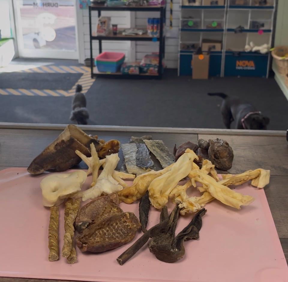 An assortment of natural dog treats and chews arranged on a pink cutting board in a pet store. In the background, two dogs are visible on a dark mat inside the store.